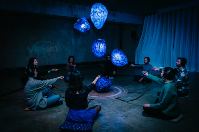 Vue du spectacle "Les Gardiens de la Montagne". Des spectateurs en cercle tendent la main vers des pierres bleues, comme en lévitation au centre de la pièce.